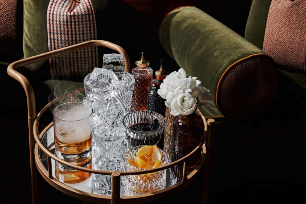 An elegant bar cart with crystal decanters, cocktail glasses, a whiskey drink with ice, and a white flower, set against a backdrop of plush green and houndstooth pillows.